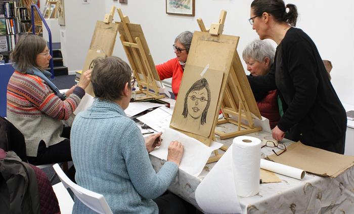 5 figures around a table with 4 seated at wooden easels with drawing boards showing hand drawn portraits. 1 figure is standing. Table is in a gallery